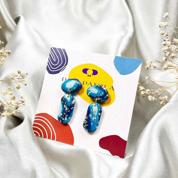 Blue earrings on a colorful card with 'Pola' branding, placed on white fabric with dried flowers.
